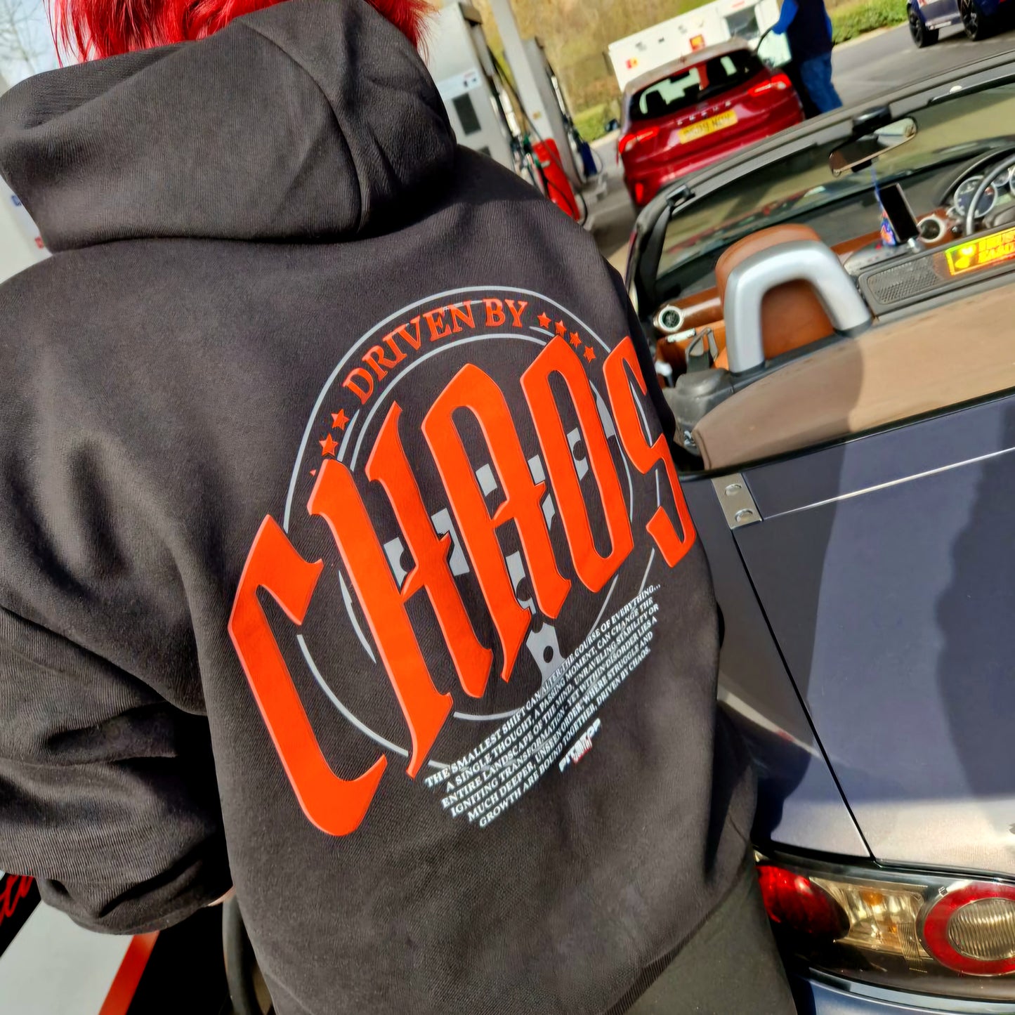 A black oversized box-fit premium hoodie with 'DRIVEN BY CHAOS' printed in large red letters within a car steering wheel on the back at Shell petrol garage getting fuel with galaxy grey mazda mx5 and JDMClare 2025