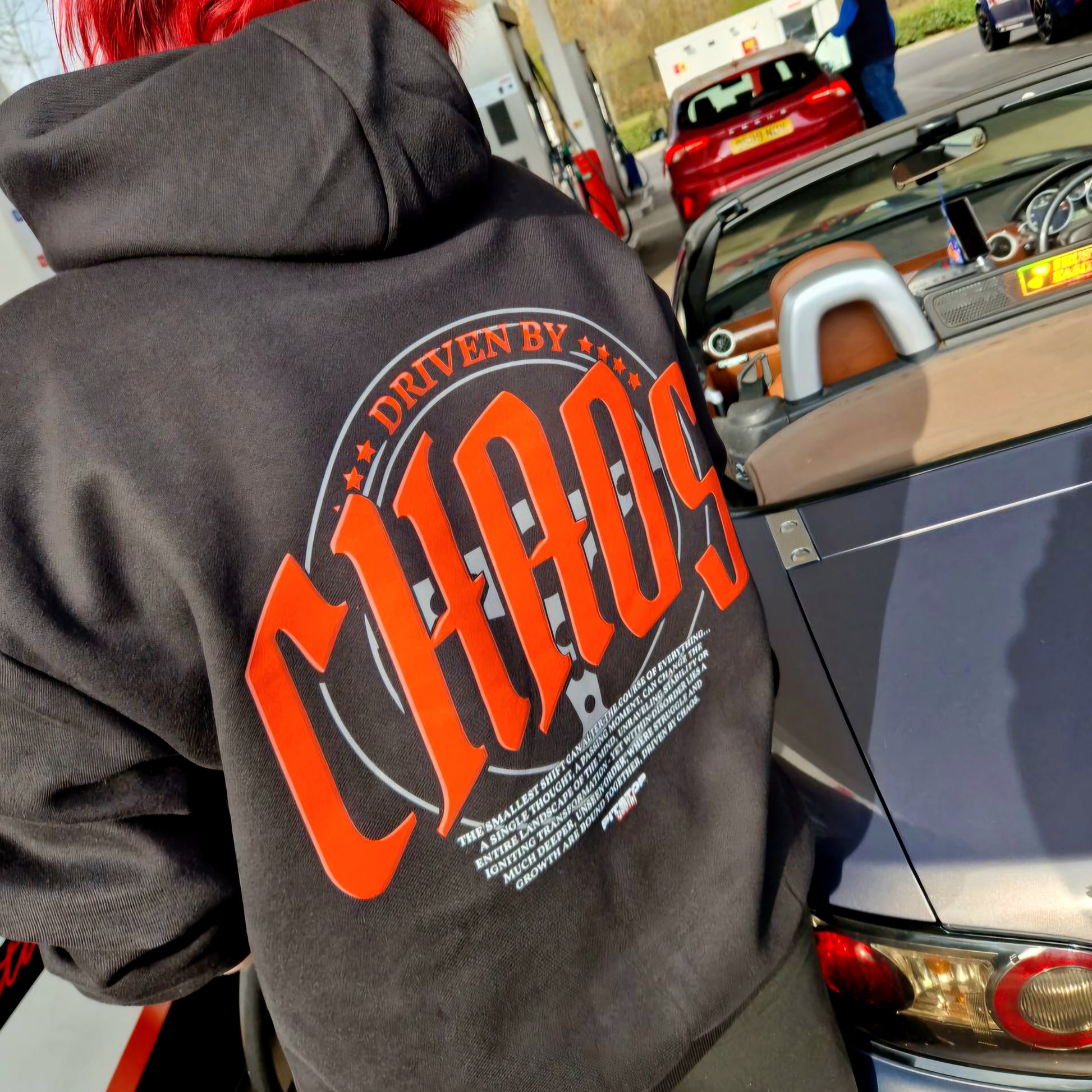 A black oversized box-fit premium hoodie with 'DRIVEN BY CHAOS' printed in large red letters within a car steering wheel on the back at Shell petrol garage getting fuel with galaxy grey mazda mx5 and JDMClare 2025