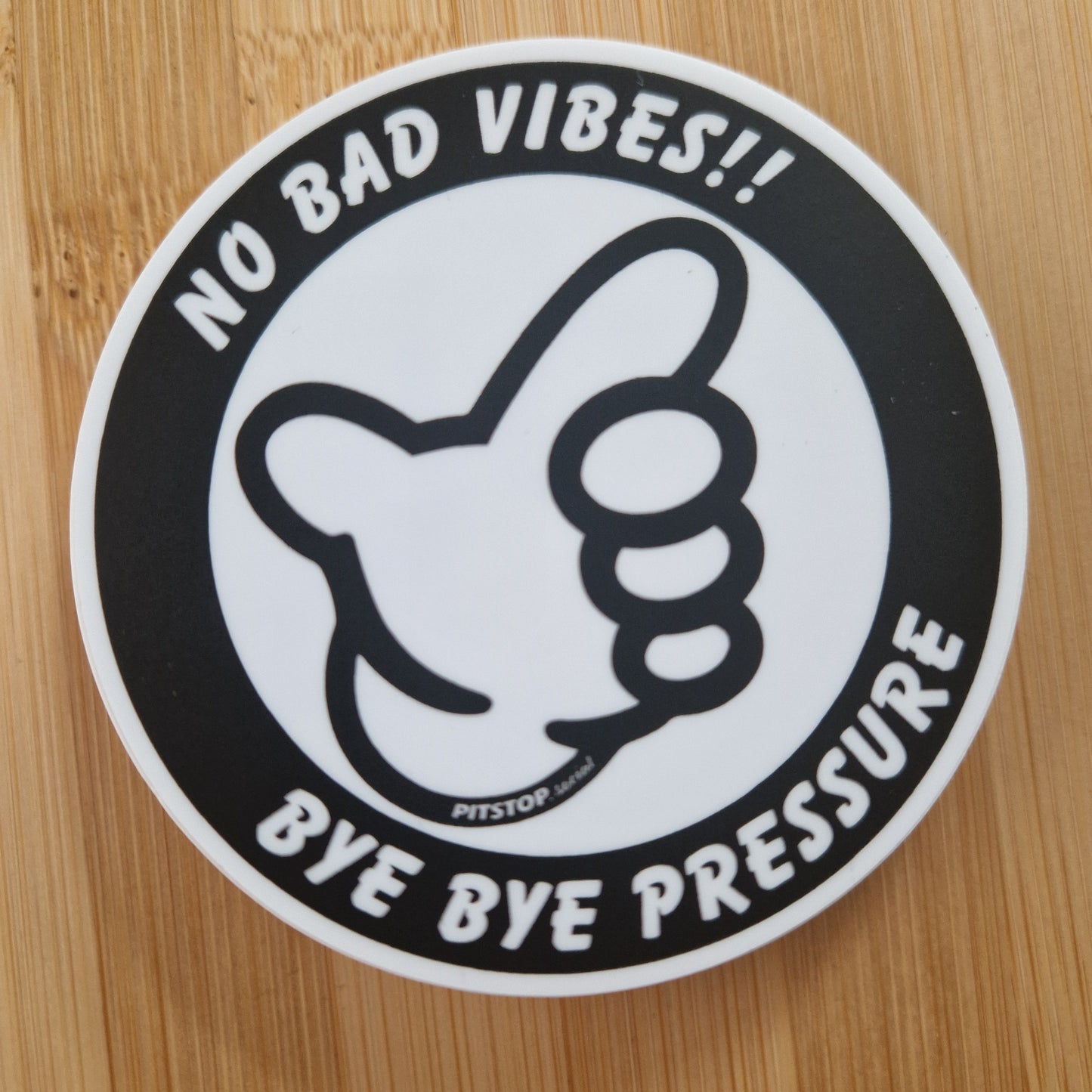 Circular sticker with a thumbs up icon and text 'No bad vibes!! Bye bye pressure' on a wooden surface.