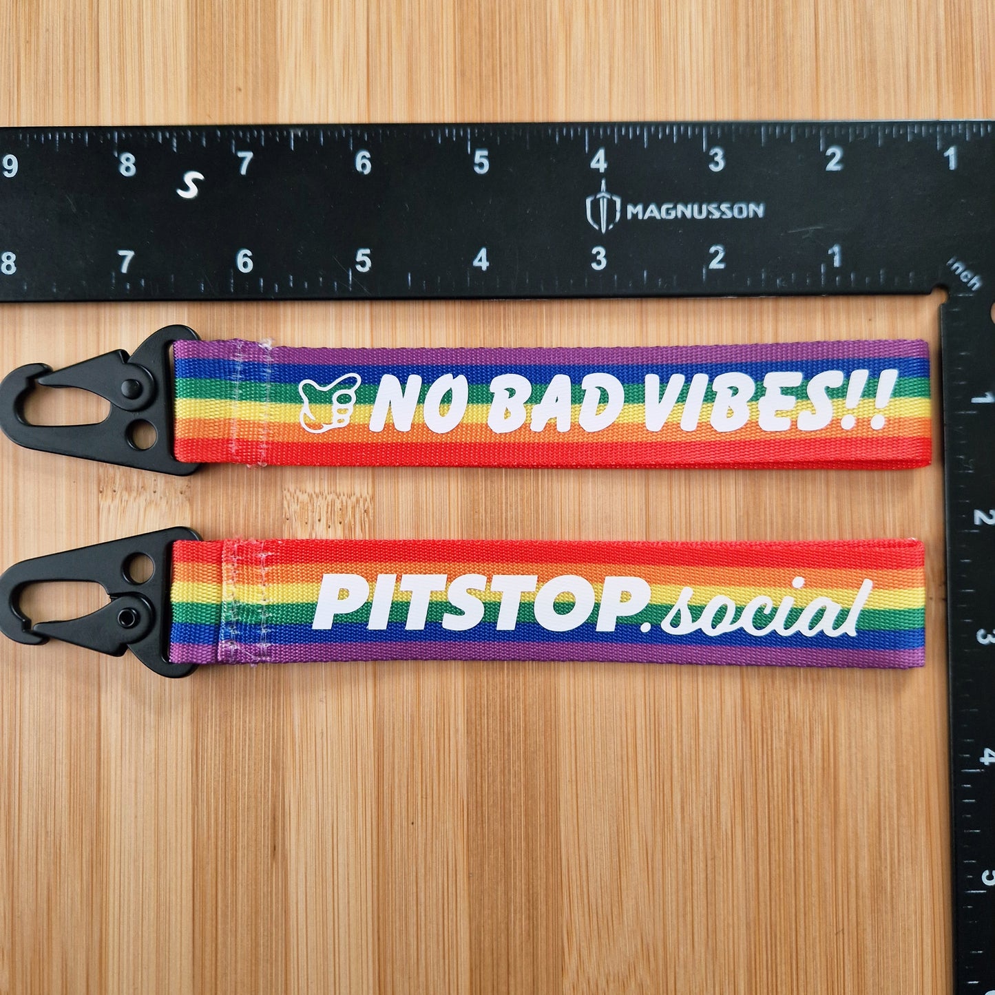 Two PITSTOP.Social branded Rainbow woven key tags with 'NO BAD VIBES' text on a wooden surface.