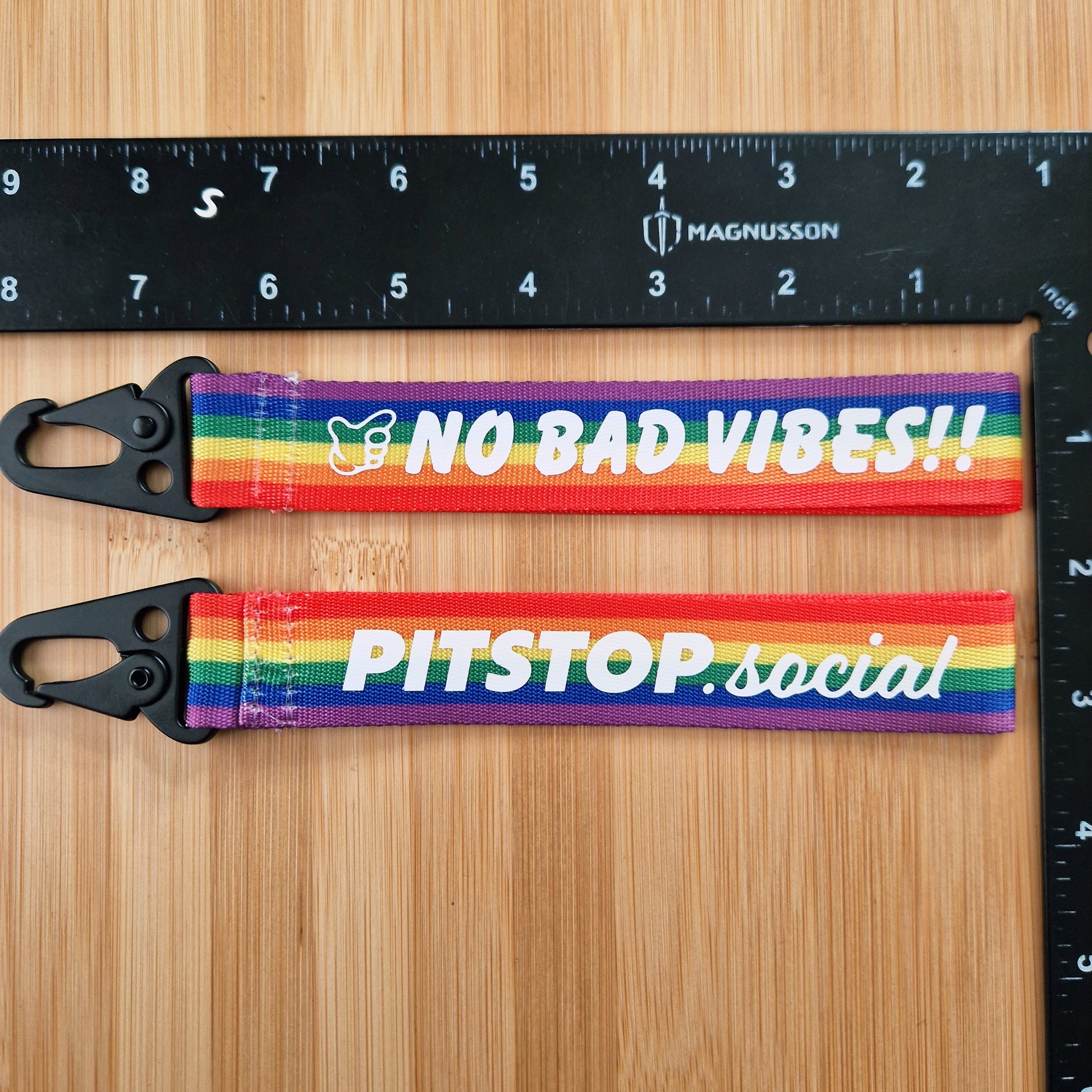 Two PITSTOP.Social branded Rainbow woven key tags with 'NO BAD VIBES' text on a wooden surface.