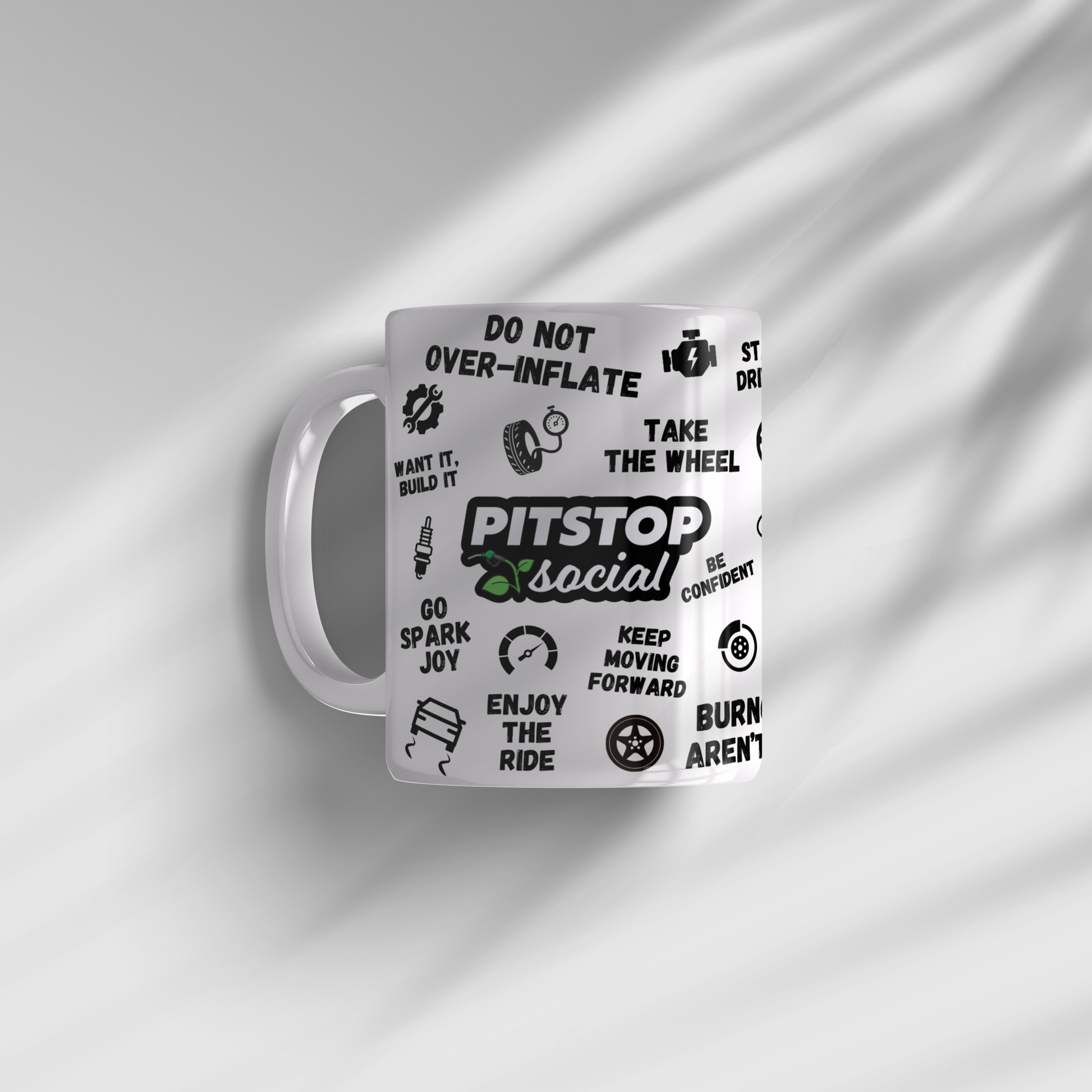 A white ceramic mug with 3D raised text and graphics including the Pitstop Social logo, various symbols of automotive relation, sunlight coming through onto plain white background.