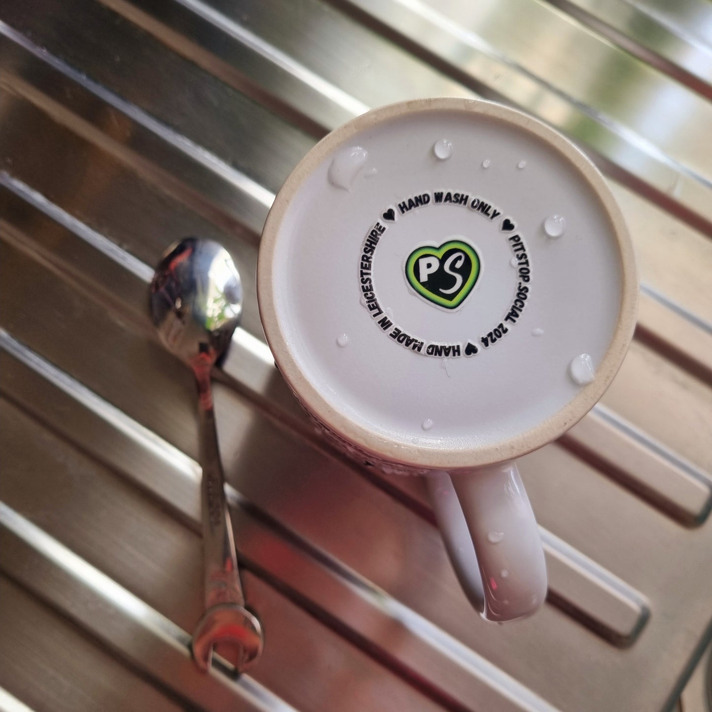 A white ceramic mug with 3D raised text and graphics including the Pitstop Social logo and text "hand made in Leicestershire" placed on a kitchen draining board.