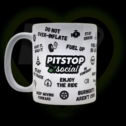 A white ceramic mug with 3D raised text and graphics including the Pitstop Social logo, various symbols, and a yellow lid with a spoon in it, placed on a wooden surface.