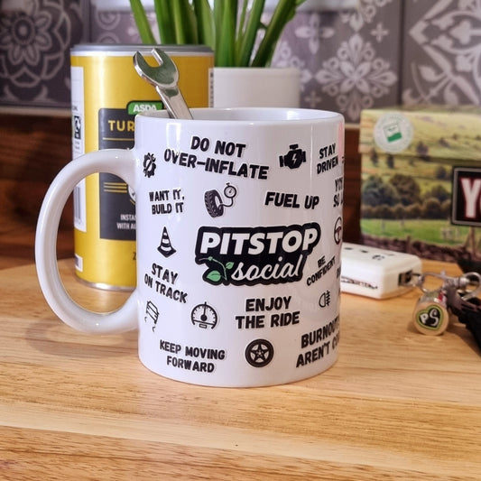 A white ceramic mug with 3D raised text and graphics including the Pitstop Social logo, various symbols, and a yellow lid with a spoon in it, placed on a wooden cutting board.