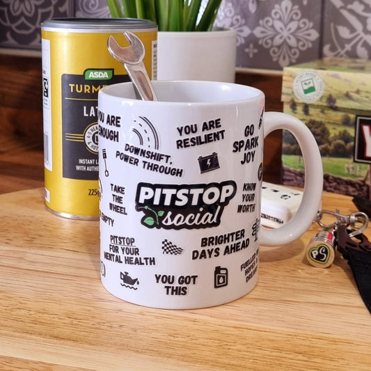 A white ceramic mug with 3D raised text and graphics including the Pitstop Social logo, various symbols, and a yellow lid with a spoon in it, placed on a wooden surface and Keys in background